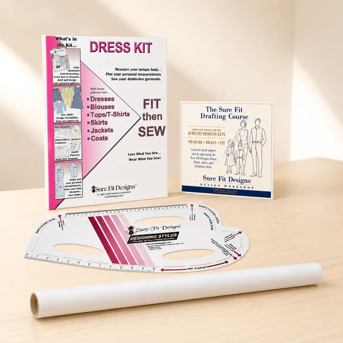 A sewing dress kit set displayed on a light wood surface, including a printed instruction booklet, a drafting course card, a curved measuring ruler, and a roll of white fabric.