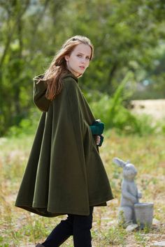 Woman in a green coat standing outdoors with trees and a stone rabbit in the background