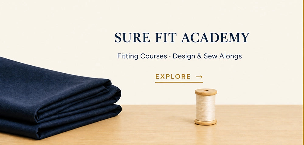 A folded navy fabric bundle sits on a light wood surface beside a spool of white thread, with centered text reading 'SURE FIT ACADEMY' and smaller text 'Fitting Courses • Design & Sew Alongs' plus an 'EXPLORE' link.