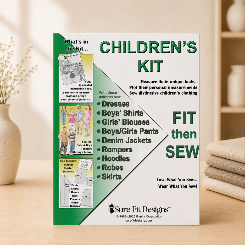 A sewing kit pattern for children's clothing by Sure Fit Designs, standing upright on a wooden surface with a soft-focus warm indoor background. The box is white and green with large text reading 'CHILDREN'S KIT' and 'FIT then SEW', plus a list of garment types and a small product photo panel on the left.