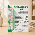 A sewing kit pattern for children's clothing by Sure Fit Designs, standing upright on a wooden surface with a soft-focus warm indoor background. The box is white and green with large text reading 'CHILDREN'S KIT' and 'FIT then SEW', plus a list of garment types and a small product photo panel on the left.