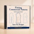 A CD case for 'Fitting Commercial Patterns' by Sure Fit Designs, featuring three garment pattern diagrams on the cover, photographed on a light wooden surface against a warm beige background.