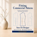 A book or booklet titled "Fitting Commercial Patterns" by Sure Fit Designs Design Workshop stands upright on a light wood surface beside a small beige vase with dried branches. The cover shows three garment pattern outlines in blue and tan on an off-white background.