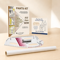 A sewing pattern kit set on a light wood surface with a warm beige background. The set includes a white pattern transfer sheet, a white roll of transfer paper, a metal ruler, and two printed instruction/booklet cards: one titled 'PANTS KIT' and the other 'The Sure Fit Drafting Course'.