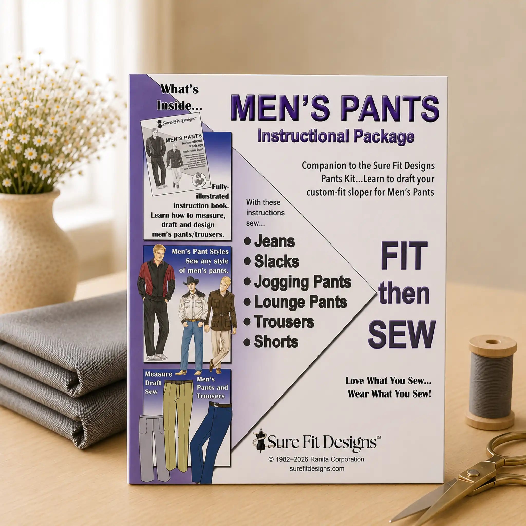 A sewing instruction booklet titled 'MEN'S PANTS Instructional Package' by Sure Fit Designs, standing upright on a tabletop with folded gray fabric, scissors, and a spool of thread nearby. The booklet cover includes photos of men's pants and text such as 'FIT then SEW'.