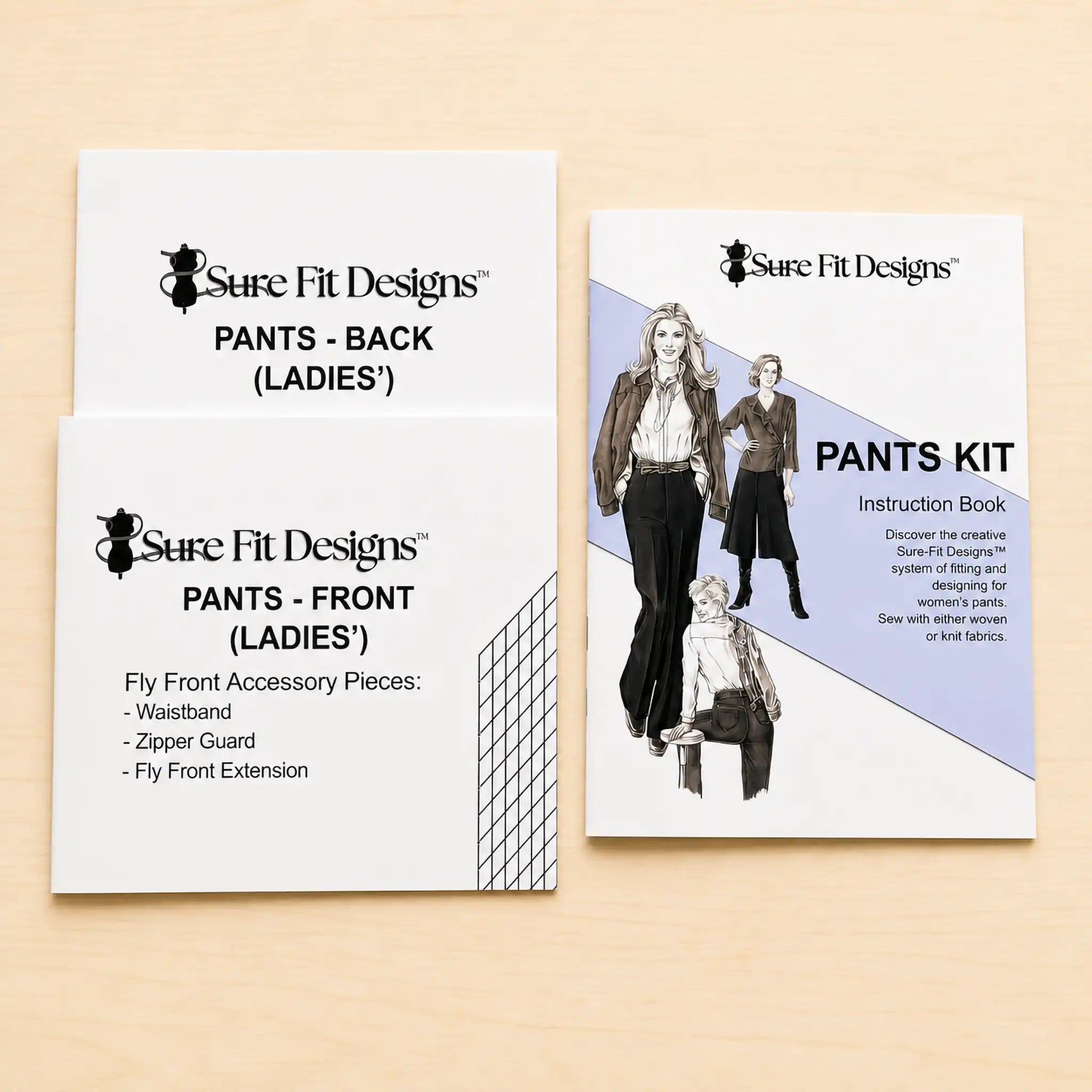 Three Sure Fit Designs sewing kit components for making women's pants laid out on a light wood surface: two white paper pieces labeled 'PANTS - FRONT (LADIES')' and 'PANTS - BACK (LADIES)' plus one instruction book titled 'PANTS KIT'.