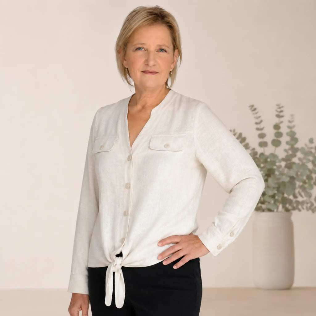 A woman modeling a light beige linen button-up shirt with a tie-front detail and chest pockets, paired with black pants, set against a warm neutral background with a potted eucalyptus plant.
