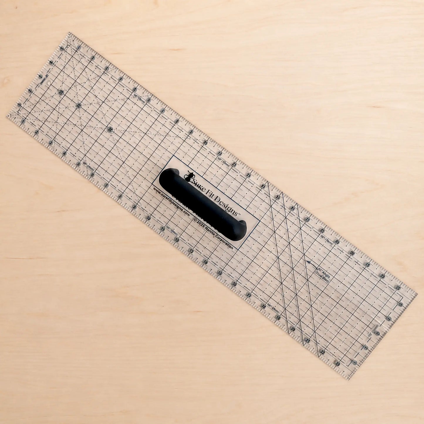 A clear acrylic rotary cutter ruler with black grid lines and a black handle, placed diagonally on a light wood background.