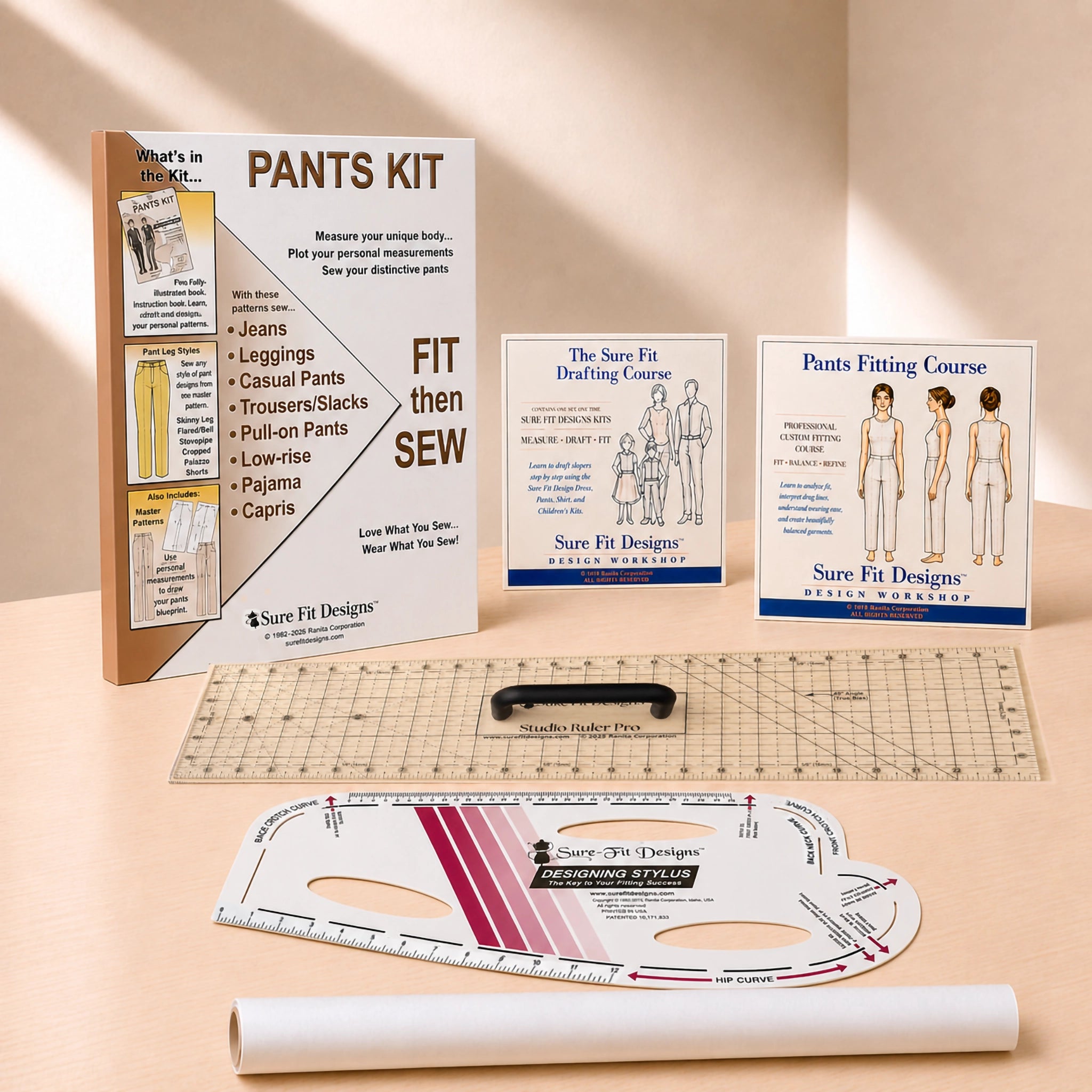 A sewing pattern kit and fitting tools arranged on a warm beige studio surface. Visible items include a 'PANTS KIT' instruction booklet, two Sure Fit Designs course cards, a clear acrylic ruler, a black sewing machine foot, a curved measuring template, and a white fabric roll.